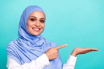 Photo of dreamy cute young lady dressed islamic hijab smiling holding arm pointing empty space isolated teal color background