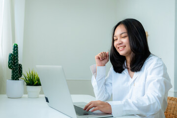 Portrait Cute Asian teen with long hair Wearing a white shirt, sitting online studying with a laptop computer on the bedroom table looks very happy.