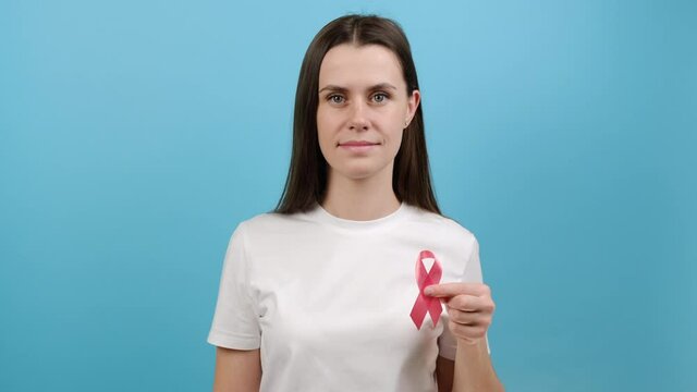 Portrait of beautiful young caucasian female 20s old years in t-shirt showing pink ribbon, model posing over blue background. Health, prevention and breast cancer awareness concept. Selective focus