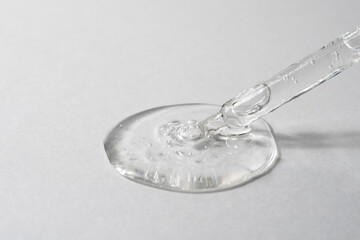 Facial serum pouring from glass pipette on grey textured background. Transparent liquid gel drop...