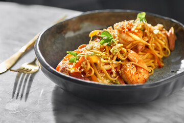 Stir fry noodles with vegetables and shrimps and seafood in black bowl on a marble table.