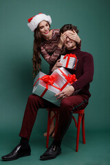 Fototapeta premium Beautiful young woman, evening makeup stylish dress , brunette, and young man in sweater, gift boxes red silk bows New Year, Christmas. Fashion photo. Green background. New year's Christmas cap