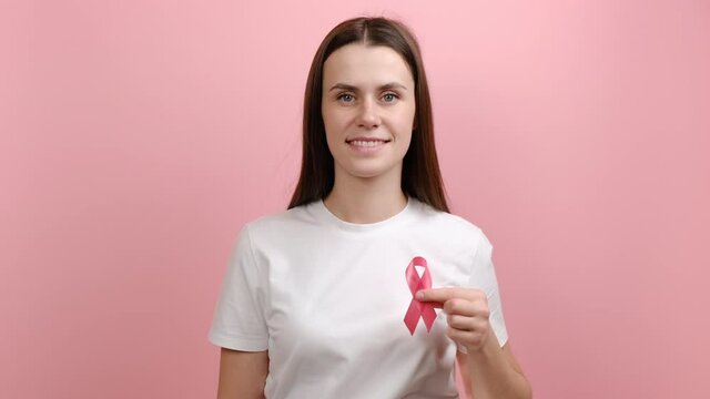 Kind cute young female holding pink ribbon, symbol of breast cancer awareness, recommendation and warning of cancer prevention, female health diagnostics, isolated over color background in studio
