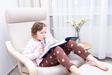 A girl, a Caucasian, sits on a soft armchair and reads a children's book.