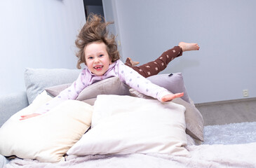 Caucasian girl cheerfully jumping on a soft sofa. The child smiles and jumps on the sofa cushions.