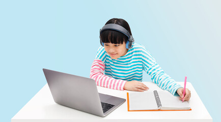 A little girl is wearing headphones and using a laptop to take notes for online learning.