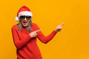 Fototapeta na wymiar Amazed gray-haired woman in santa hat and sunglasses standing isolated over yellow background pointing with fingers on the blank space for advertisement, mockup image, check this out, funny concept