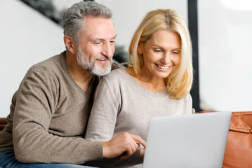 Lovely mature spouses using laptop computer for spending leisure at home, watching movie together, choosing purchases, shopping. Middle-aged couple sits on the sofa and watching at the screen
