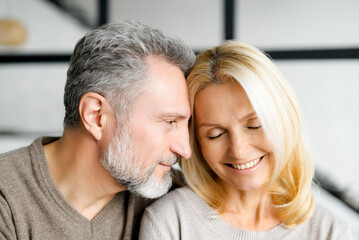 Headshot of romantic middle-aged couple indoors. Handsome grey-haired mature man watching with passion and love at beautiful charming mid-age woman. Mature spouses at home