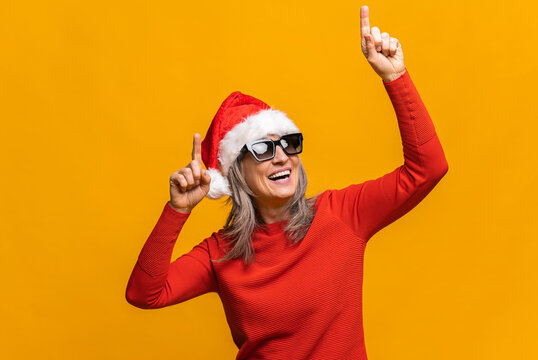 Mature Lady In Santa Hat And Sunglasses Dancing Active Isolated On Yellow, Joyful Modern Senior Woman With Gray-hair Enjoying Celebrating New Year Party