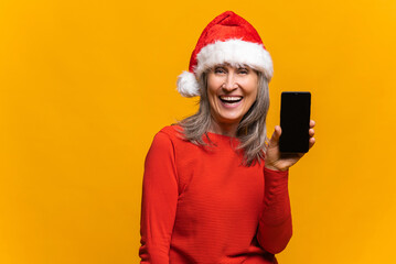 Delighted by the holiday deal mature woman in santa hat holding smartphone with empty screen, overjoyed middle-aged lady demonstrates phone with blank display, isolated on yellow