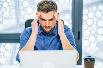 A businessman sits in front of a laptop, touches his temples, feels stress, problems in business. Male office worker at workplace feels overwhelmed and suffers from throbbing headache