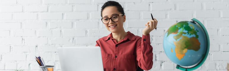 happy teacher in eyeglasses using laptop and holding pen while working from home, banner. © LIGHTFIELD STUDIOS