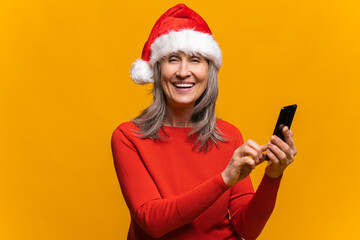 Close-up portrait of hilarious senior woman in santa hat using smartphone isolated on yellow, middle-aged gray-haired lady sending New Year congratulations, texting online