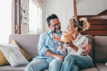 Happy caucasian couple holding piggy bank to save money to make their future dreams come true. Loving man and woman holding piggy bank for savings and discussing while sitting on sofa at home