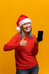 Vertical portrait of hilarious senior woman in red santa hat holding smartphone with empty screen isolated on yellow, middle-aged gray-haired lady showing thumb up, like sign