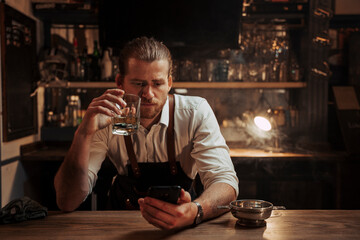 Caucasian male bartender working alone having a drink while texting on cellular device 