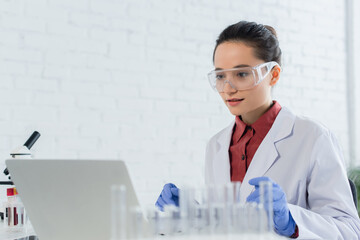 young scientist in white coat and goggles looking at laptop near test tubes.