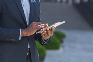 close up of afro american businessman hands using tablet. A black business man in a formal suit browses the internet mobile device. Closeup. outside outdoors. Urban modern office building background