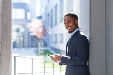 Fototapeta premium Successful and happy african american businessman near office holding mobile phone smiles and rejoices reads news and writes