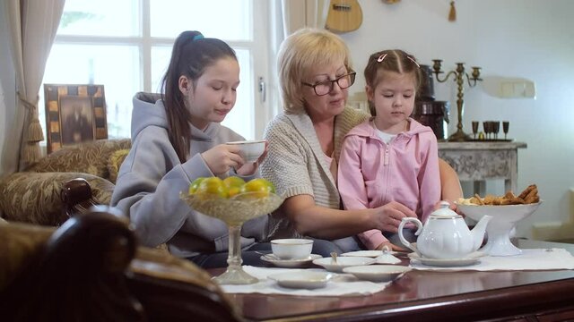Grandmother and granddaughters are drinking tea. Two sisters visiting a caring grandmother. A family friendly environment for sweet treats.
