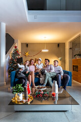 A happy family is sitting on the couch in living room and having a video call via tablet. The room is decorated in the Christmas spirit. Also, their dog is on the couch with them.