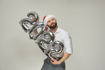 A smiling man in a red velvet Santa hat is hugging silver balloons in the shape of 2022 gently. A...