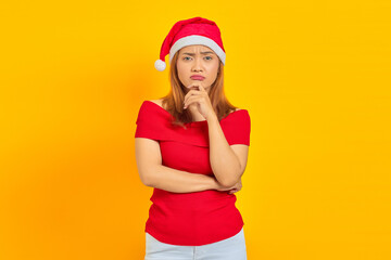 Pensive young woman wearing Christmas hat thinking about something on yellow background