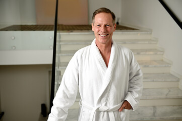 Portrait of a confident handsome middle aged man wearing terry bathrobe and smiling toothy smile looking at camera standing on the stairs of health spa while enjoying relaxing male beauty procedures