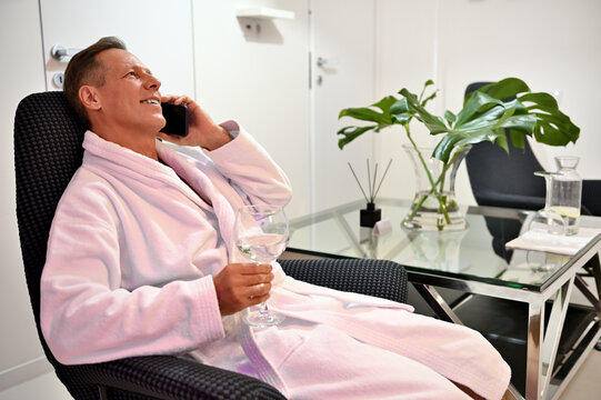 Relaxed handsome middle aged 50 years old man in white terry bathrobe sitting on an armchair with a glass of fresh water and talking on mobile phone at wellness spa center