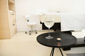 Focus on a glass of mineral water and a candle on a black round table in a white interior of a private resting room in a luxury spa wellness clinic. Aromatherapy, professional body care concept