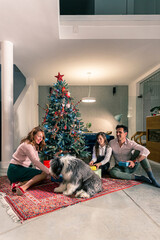 A mother and father sitting with their daughter on the floor next to the Christmas tree and exchanging Christmas presents. The living room is decorated in the spirit of the upcoming holidays.