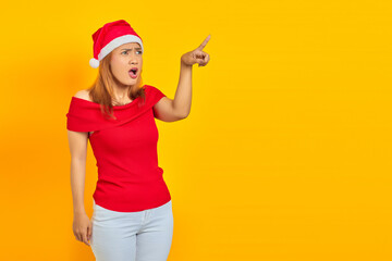 Surprised young Asian woman wearing Christmas hat and pointing sideways over yellow background