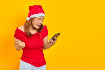 Cheerful young Asian woman wearing Christmas hat holding mobile phone and celebrating success on yellow background