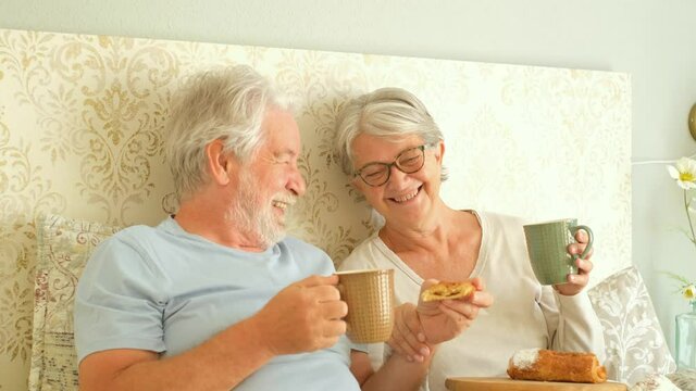 Old senior caucasian couple enjoying breakfast in the morning at bed in the bedroom at home. Elderly couple eating croissant and drinking coffee from cup for breakfast at home.
