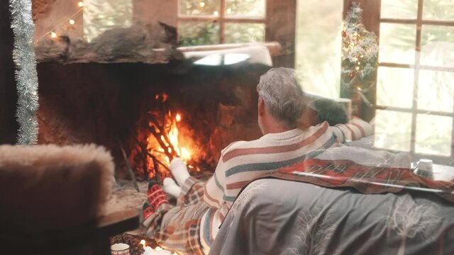 Old caucasian couple spending leisure time together at home. Loving romantic husband and wife relaxing while looking at burning fireplace during winter christmas holiday
