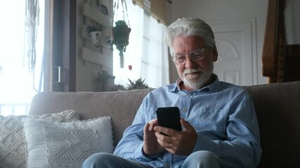 One old man using phone at home sitting on the sofa smiling and having fun learning chatting with friends and surfing on the web. Satisfied male senior indoor with smartphone.

