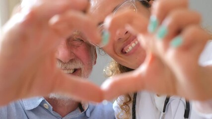 Close up of young female doctor and senior man showing heart sign with hand at home. Doctor helping senior patient and giving care. Elderly medical health care.
