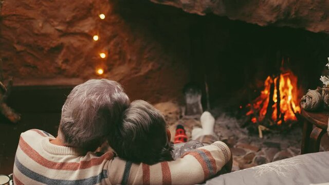 Old caucasian couple spending leisure time together at home. Loving romantic husband and wife relaxing while looking at burning fireplace during winter christmas holiday
