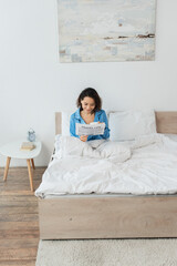 cheerful young woman in pajamas reading travel life newspaper and holding cup in bed.