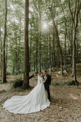 The groom in a black suit and the bride in a white dress stand and hug each other in the green forest