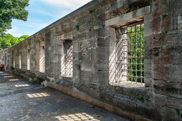 A row of antique barred windows in the shade. Ancient wall of gray stone. Historical sights and tourist locations.