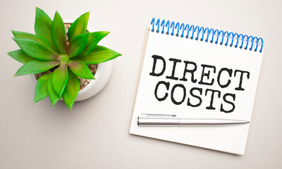 DIRECT COSTS is written in a white notepad near a clipboard, calculator, green plant, glasses and a pen on a yellow and concrete background. Business concept. Flat lay.