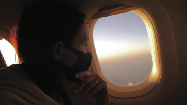 Young woman sits on a plane in a black medical respiratory mask and looks out the window. Health, travel concepts. Safety and new rules of conduct for air transport passengers.