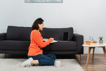 woman sitting on carpet and writing in notebook near laptop with blank screen on couch.