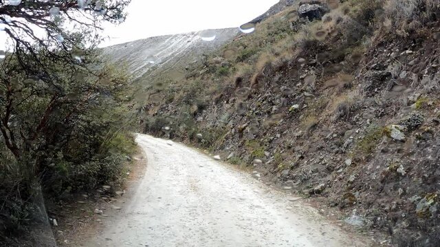 pov to the Paron lagoon, Huaras by car on the dirt road between trees and hills in the day with blue sky and white clouds and raining with drops on the windshield and using the windshield wiper