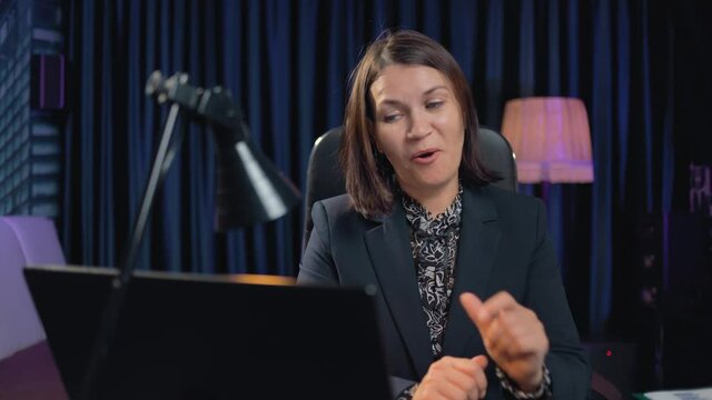 A business woman in a strict suit communicates with friends via video on a laptop. She waves her hand affably and has positive emotions on her face. The lady is at home on a remote job.