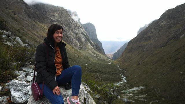 a beautiful woman sitting on a stone resting from climbing a hill next to a canyon in the Paron lagoon, Huaras speaking to the camera and smiling and waving