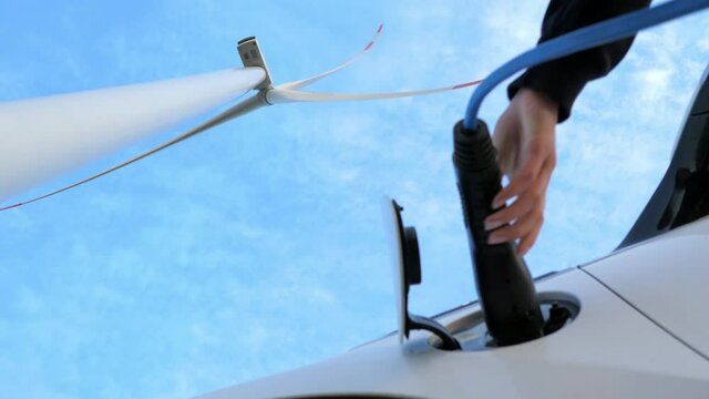 Windmill generates electricity against blue sky. Young woman disconnects plug off white car and closes cap under rotating propeller low angle shot