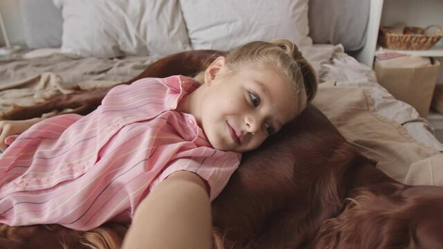 Handheld POV shot of pretty 8-year-old girl recording video of herself talking while lying on cute Irish setter dog resting together on bed in cozy bedroom